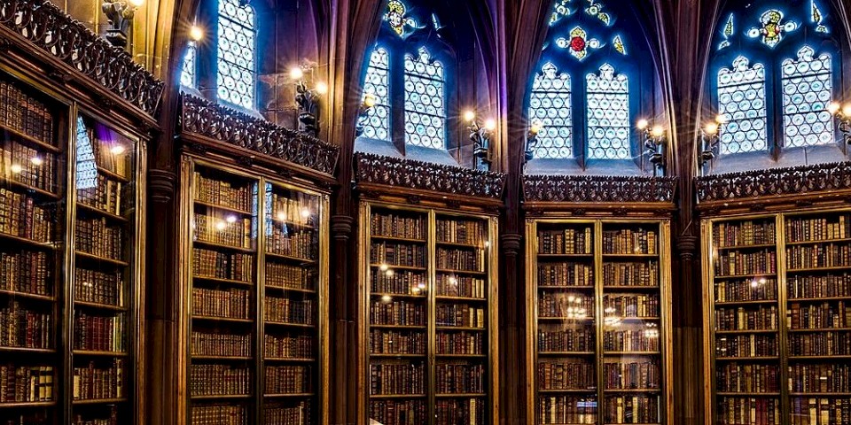 Bild zeigt alte Bücher in hohen Bücherregalen. Licht fällt durch gotische Fenster hinein. (John Rylands Library Manchester)