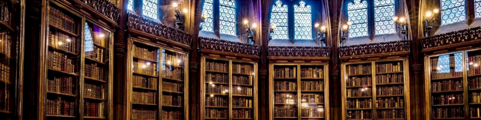 Bild zeigt alte Bücher in hohen Bücherregalen. Licht fällt durch gotische Fenster hinein. (John Rylands Library Manchester)