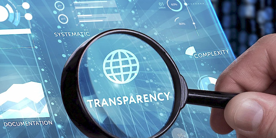 Hand with magnifying glass examining the value ‘Transparency’ on a digital screen display – symbolic of scientific work and transparency