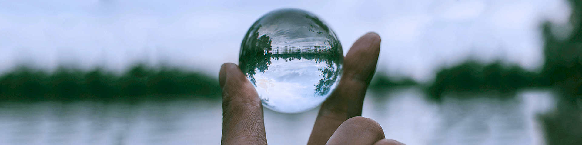 Hand holding a glass ball between two fingers in front of a lake, in which the lake landscape is reflected horizontally – symbolic illustration of the phenomenological perspective of homeopathic medicine prescription.