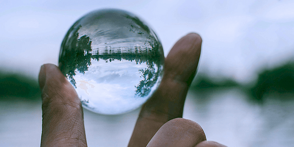 Hand holding a glass ball between two fingers in front of a lake, in which the lake landscape is reflected horizontally – symbolic illustration of the phenomenological perspective of homeopathic medicine prescription.