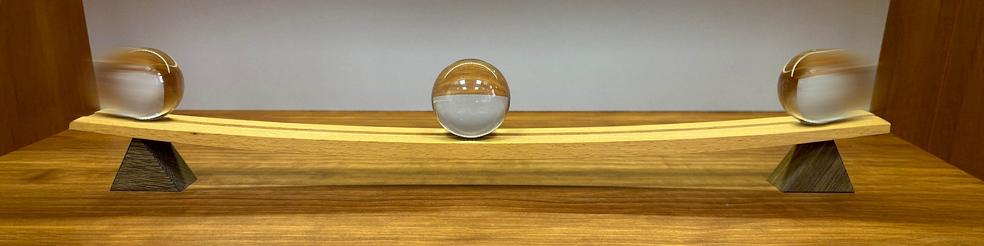Symmetrically curved wooden track with a glass ball resting in the centre, and a glass ball on the left and right, each moving towards the centre, symbolising the principles of dialectics.