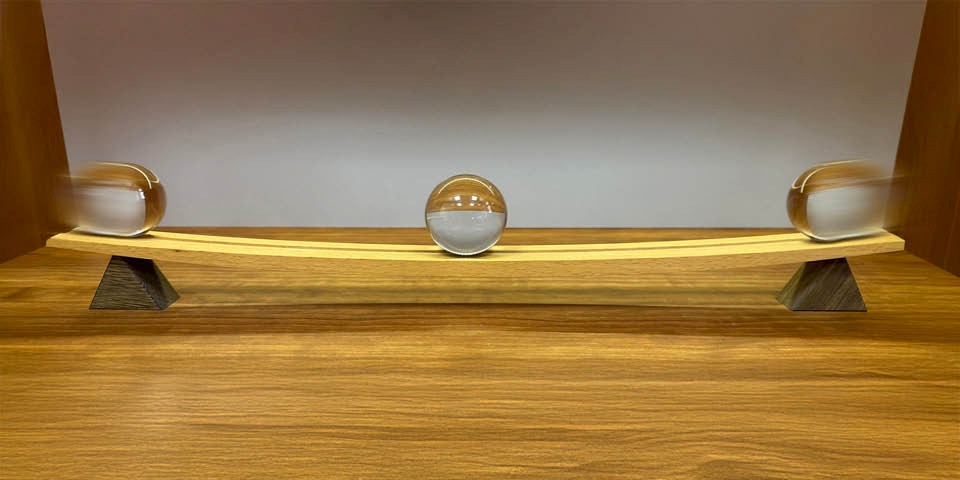 Symmetrically curved wooden track with a glass ball resting in the centre, and a glass ball on the left and right, each moving towards the centre, symbolising the principles of dialectics.