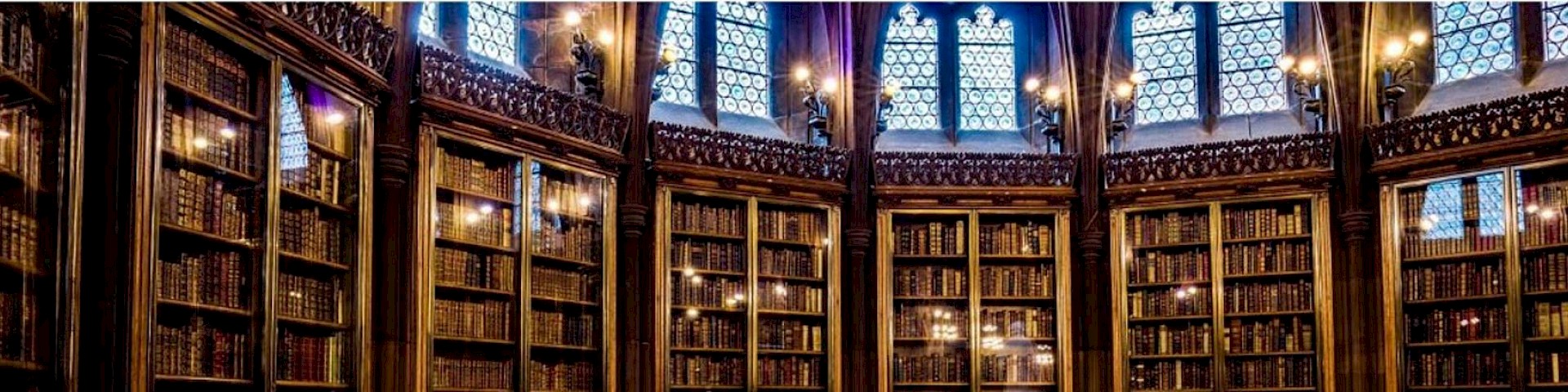 Picture shows old books on high bookshelves. Light falls in through gothic windows. (John Rylands Library Manchester)